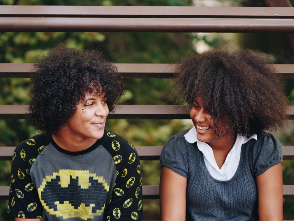 two young people smiling at eachother while sitting on a bench outside